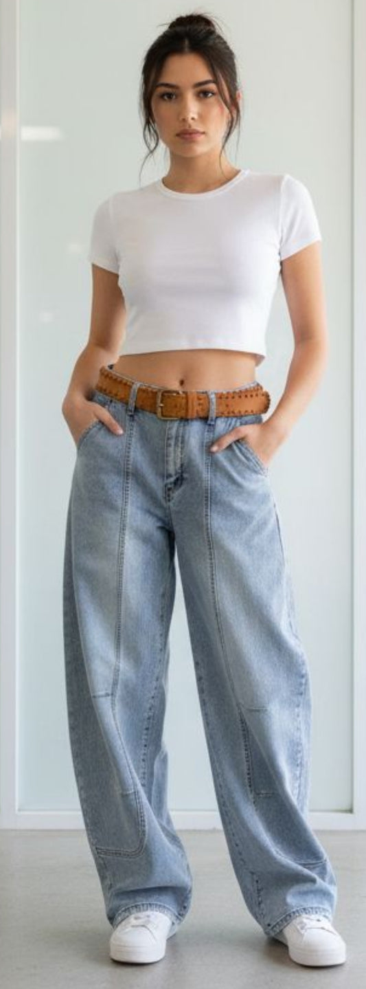 Damen trägt hellblaue High Waist Baggy Jeans mit weitem Bein, Herz-Taschen hinten und braunem Gürtel, kombiniert mit weißem Crop Top und Sneakern.