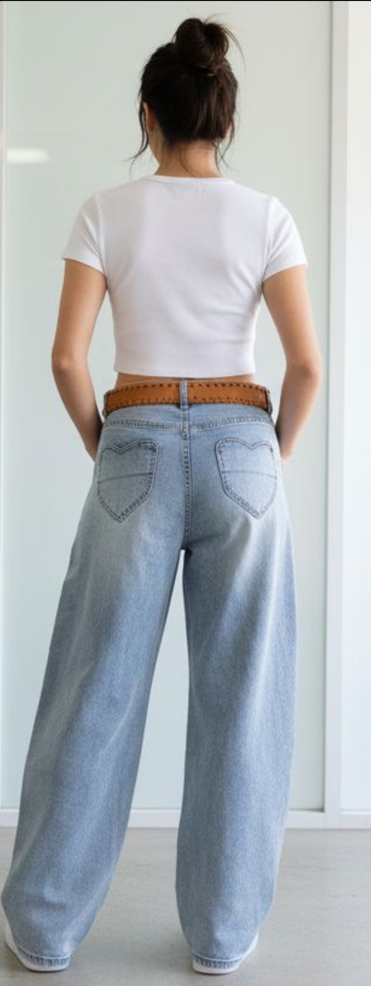 Damen trägt hellblaue High Waist Baggy Jeans mit weitem Bein, Herz-Taschen hinten und braunem Gürtel, kombiniert mit weißem Crop Top und Sneakern.